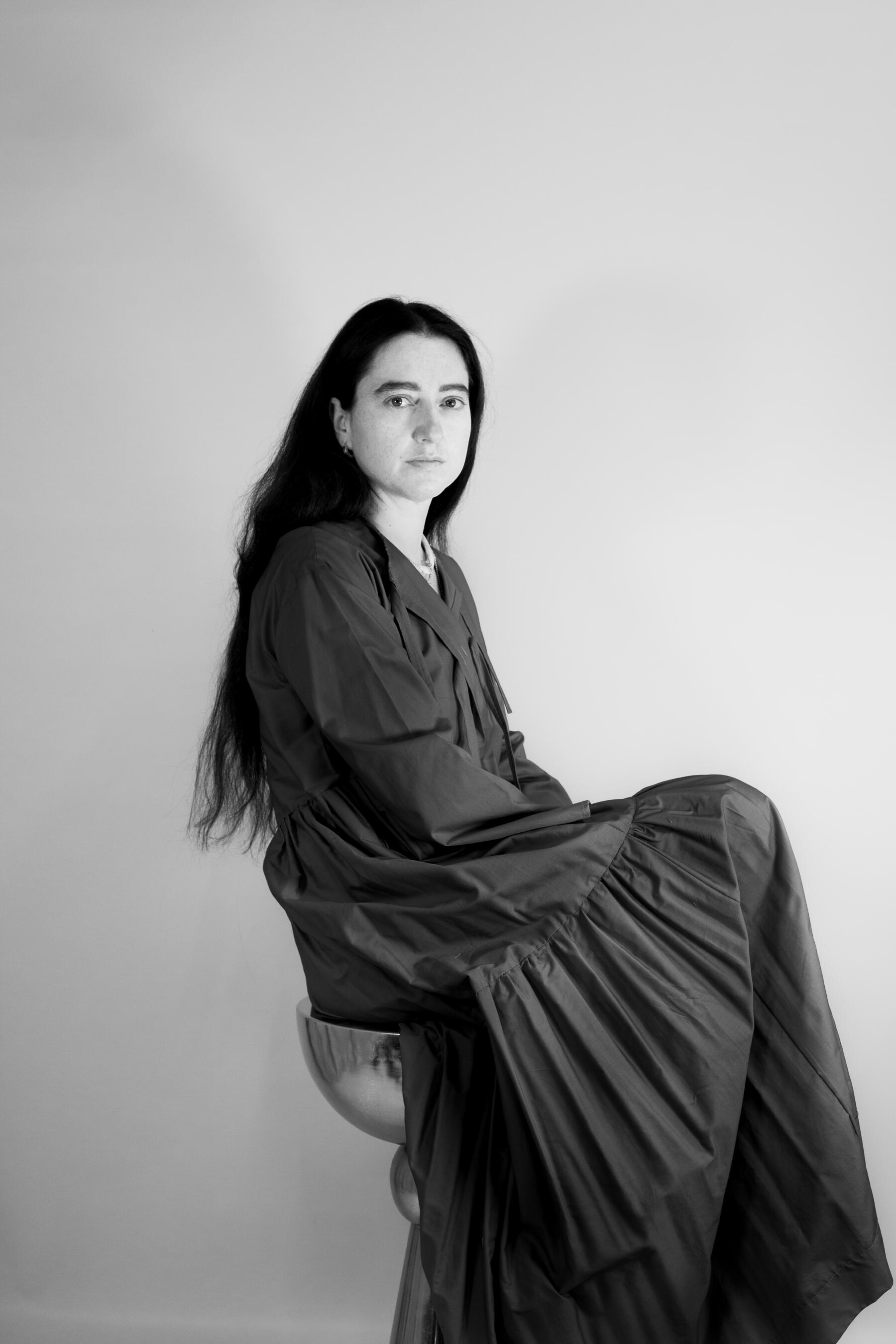 Alina Stanila | British Fashion Designer & Founder of Mystic Moods Black-and-white seated portrait of Alina Stanila, British fashion designer and founder and creative director of Mystic Moods, wearing a long dark dress.